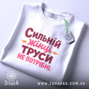 Футболка труси не потрібні. Біла футболка, складена на поверхні, з принтом 'Сильний жінці труси не потрібні' в червоному і рожевому кольорах, з жовтими елементами. На футболці є етикетка з зеленим шнурком. Фон світло-сірий.