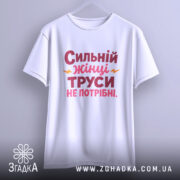Футболка труси не потрібні. Біла футболка, підвішена на вішалці, з принтом 'Сильний жінці труси не потрібні' в червоному і рожевому кольорах, з жовтими акцентами. Фон світло-сірий.
