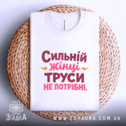 Футболка труси не потрібні. Біла футболка, складена на плетеній кошику, з принтом 'Сильний жінці труси не потрібні' в червоному і рожевому кольорах, з жовтими елементами. Фон світло-сірий.