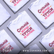 Футболка труси не потрібні. Біла футболка з принтом на грудях, на якому написано 'Сильний жінці труси не потрібні' червоним і рожевим кольорами, з жовтими акцентами. Модель позує з легким усмішкою, фон світло-сірий.