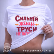 Футболка труси не потрібні. Біла футболка, складена на поверхні, з принтом 'Сильний жінці труси не потрібні' в червоному і рожевому кольорах, з жовтими елементами. На футболці є етикетка з зеленим шнурком. Фон світло-сірий.