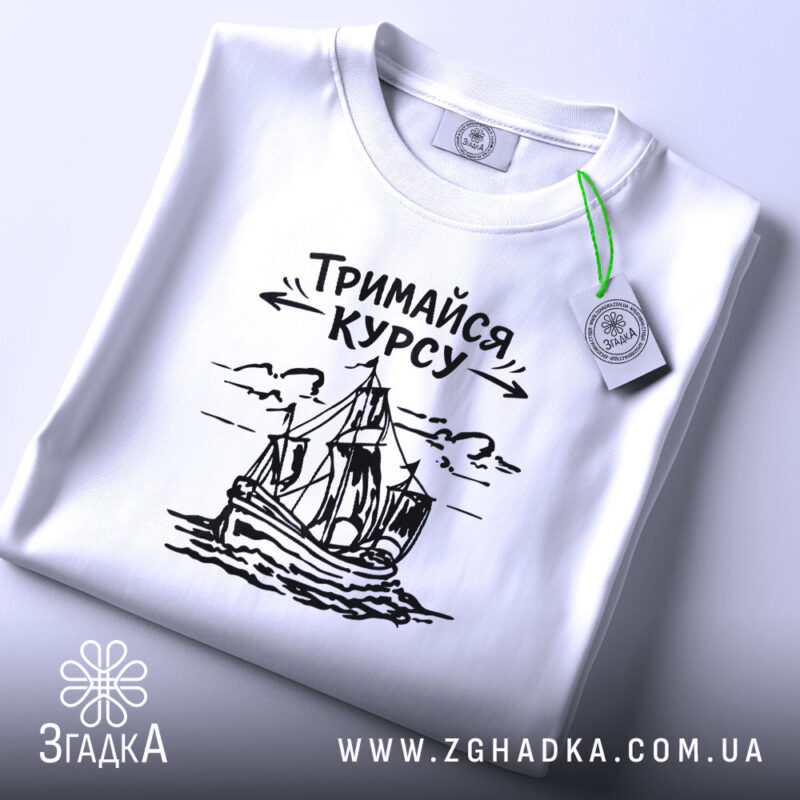 Футболка тримайся курсу, біла, лежить на плоскій поверхні, фон світло-сірий, принт з малюнком корабля та написом 'Тримайся курсу' у чорному кольорі, на комірці є етикетка.