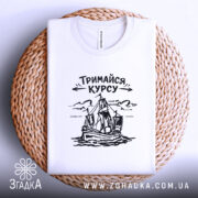Футболка тримайся курсу, біла, складена на плоскій поверхні, фон світло-сірий, принт з малюнком корабля та написом 'Тримайся курсу' у чорному кольорі, лежить на плетеній основі.