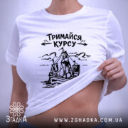 Футболка тримайся курсу, біла, лежить на плоскій поверхні, фон світло-сірий, принт з малюнком корабля та написом 'Тримайся курсу' у чорному кольорі, на комірці є етикетка.