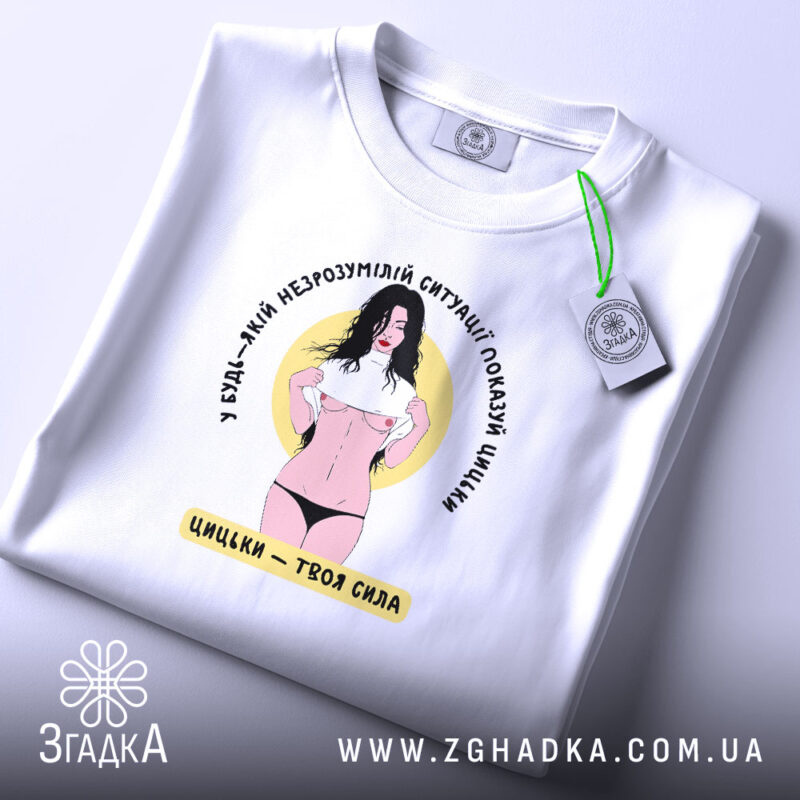 Футболка цицьки білого кольору, зображена в плоскому вигляді, фон світло-сірий, принт з жінкою в чорному нижній білизні, яка піднімає футболку, текст навколо принта написаний чорним кольором, на етикетці зелена стрічка.
