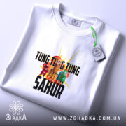 Футболка Tung Tung Sahur білого кольору, складена на поверхні, з видимим принтом, фон світло-сірий, на етикетці присутня зелена стрічка.