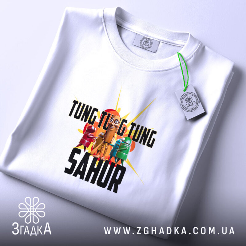 Футболка Tung Tung Sahur білого кольору, складена на поверхні, з видимим принтом, фон світло-сірий, на етикетці присутня зелена стрічка.