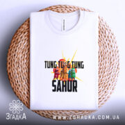 Футболка Tung Tung Sahur білого кольору, складена на плетеній кошику, фон світло-сірий, принт з персонажами та текстом чітко видимий на поверхні.