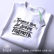 Футболка тусуюся, біла, складена на поверхні, фон світло-сірий, принт чорний з текстом 'Тусуюся там, де добре годують.', з етикеткою зеленого кольору.