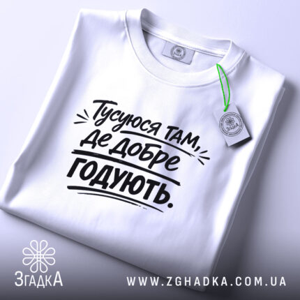 Футболка тусуюся, біла, складена на поверхні, фон світло-сірий, принт чорний з текстом 'Тусуюся там, де добре годують.', з етикеткою зеленого кольору.