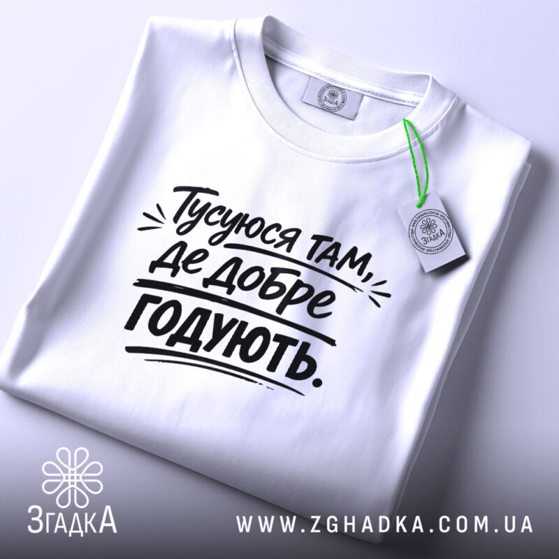 Футболка тусуюся, біла, складена на поверхні, фон світло-сірий, принт чорний з текстом 'Тусуюся там, де добре годують.', з етикеткою зеленого кольору.