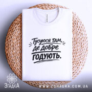 Футболка тусуюся, біла, складена на плетеній основі, фон світло-сірий, принт чорний з текстом 'Тусуюся там, де добре годують.'