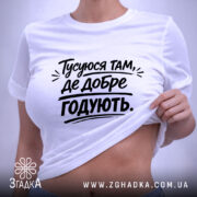 Футболка тусуюся, біла, складена на поверхні, фон світло-сірий, принт чорний з текстом 'Тусуюся там, де добре годують.', з етикеткою зеленого кольору.