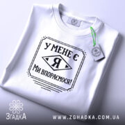 Футболка у мене є я, біла, складена на поверхні, фон світло-сірий, принт чорного кольору з написом 'У МЕНЕ Є Я МИ ВПОРЕМОСЯ!', з етикеткою зеленого кольору.