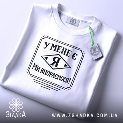 Футболка у мене є я, біла, складена на поверхні, фон світло-сірий, принт чорного кольору з написом 'У МЕНЕ Є Я МИ ВПОРЕМОСЯ!', з етикеткою зеленого кольору.