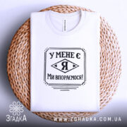 Футболка у мене є я, біла, складена на кошику з плетеного матеріалу, фон білий, принт чорного кольору з написом 'У МЕНЕ Є Я МИ ВПОРЕМОСЯ!'.