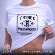 Футболка у мене є я, біла, складена на поверхні, фон світло-сірий, принт чорного кольору з написом 'У МЕНЕ Є Я МИ ВПОРЕМОСЯ!', з етикеткою зеленого кольору.