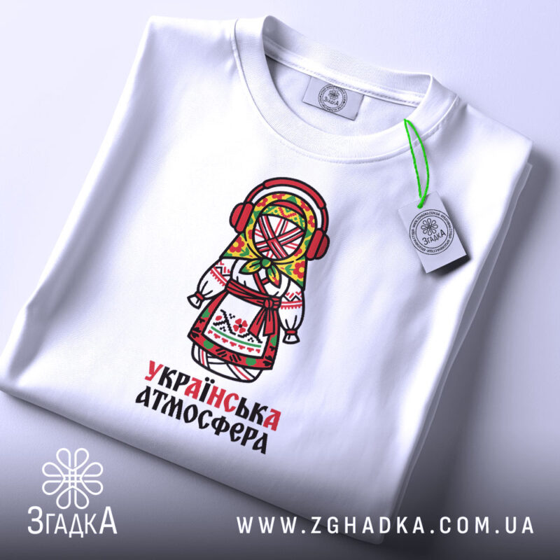 Футболка українська атмосфера, біла, на столі, з принтом ляльки-мотанки, фон світлий, деталі принта яскраві, з червоними навушниками та кольоровим головним убором, етикетка з зеленим шнурком.