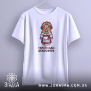 Футболка українська атмосфера, біла, підвішена на вішалці, з принтом ляльки-мотанки, фон світло-сірий, деталі принта яскраві, з червоними навушниками та кольоровим головним убором.