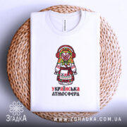 Футболка українська атмосфера, біла, на плетеній кошику, з принтом ляльки-мотанки, фон світлий, деталі принта яскраві, з червоними навушниками та кольоровим головним убором.