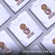 Футболка українська атмосфера, біла, на моделі, з принтом у вигляді ляльки-мотанки в традиційному вбранні, фон світло-фіолетовий, деталі принта яскраві, з червоними навушниками та кольоровим головним убором.