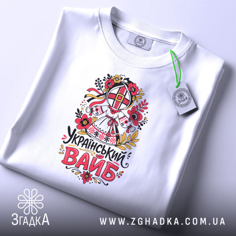 Футболка український вайб білого кольору, зображена на столі в горизонтальному положенні, фон світло-фіолетовий, принт з традиційним українським мотивом, що включає в себе квіти та ляльку-мотанку, текст "Український вайб" виконаний у яскравих кольорах, на футболці прикріплений етикет.
