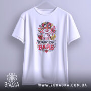 Футболка український вайб білого кольору, зображена в вертикальному положенні, фон світло-сірий, принт з традиційним українським мотивом, що включає в себе квіти та ляльку-мотанку, текст "Український вайб" виконаний у яскравих кольорах.