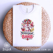 Футболка український вайб білого кольору, зображена на плетеній кошику, фон світло-фіолетовий, принт з традиційним українським мотивом, що включає в себе квіти та ляльку-мотанку, текст "Український вайб" виконаний у яскравих кольорах.