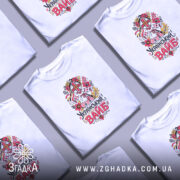 Футболка український вайб білого кольору, зображена на молодому чоловіку в анфас, фон світло-фіолетовий, принт з традиційним українським мотивом, що включає в себе квіти та ляльку-мотанку, текст "Український вайб" виконаний у яскравих кольорах.