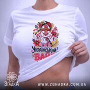 Футболка український вайб білого кольору, зображена на столі в горизонтальному положенні, фон світло-фіолетовий, принт з традиційним українським мотивом, що включає в себе квіти та ляльку-мотанку, текст "Український вайб" виконаний у яскравих кольорах, на футболці прикріплений етикет.