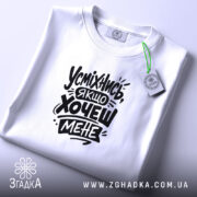 Футболка усміхнись якщо хочеш мене. Біла футболка, складена на столі, з видимим принтом чорного кольору. Фон світло-сірий, на футболці зелена етикетка.