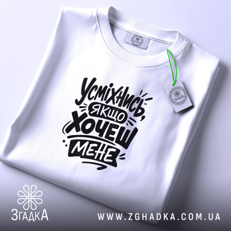 Футболка усміхнись якщо хочеш мене. Біла футболка, складена на столі, з видимим принтом чорного кольору. Фон світло-сірий, на футболці зелена етикетка.