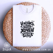 Футболка усміхнись якщо хочеш мене. Біла футболка, складена на плетеній кошику, з принтом чорного кольору. Фон світло-сірий, видима етикетка на горловині.