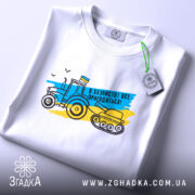 Футболка в хазяйстві все пригодиться. Біла футболка, зображена зверху, фон світло-сірий. Принт з трактором і танком у жовто-блакитних кольорах, текст "В хазяйстві все пригодиться!", етикетка з зеленим шнурком.