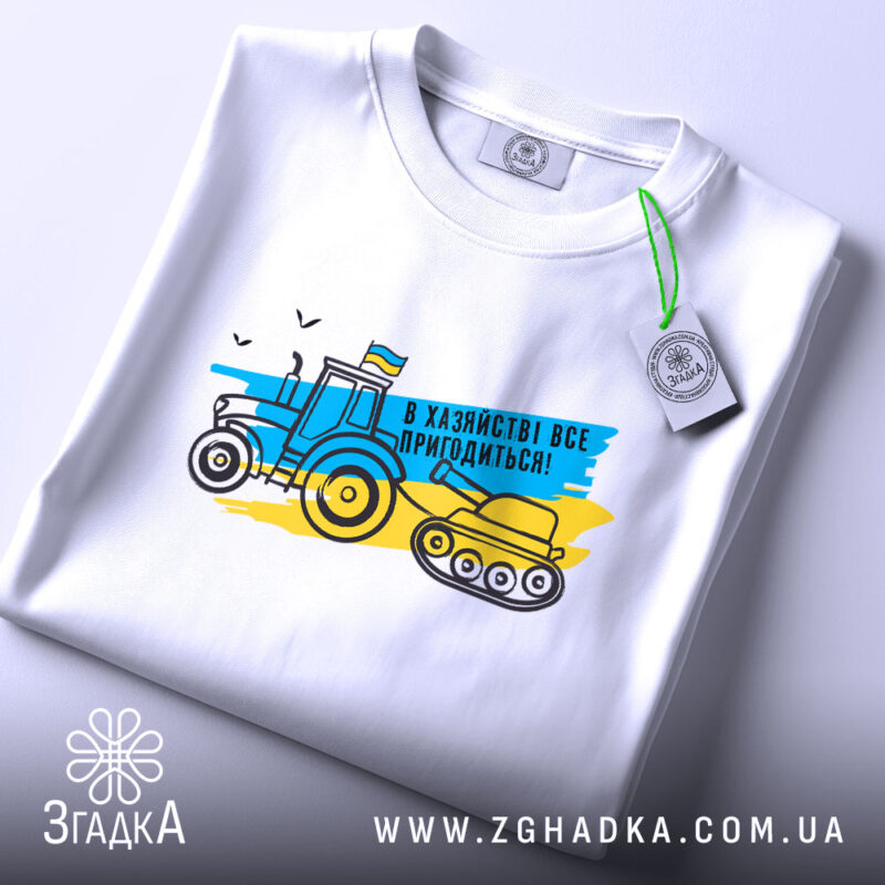Футболка в хазяйстві все пригодиться. Біла футболка, зображена зверху, фон світло-сірий. Принт з трактором і танком у жовто-блакитних кольорах, текст "В хазяйстві все пригодиться!", етикетка з зеленим шнурком.