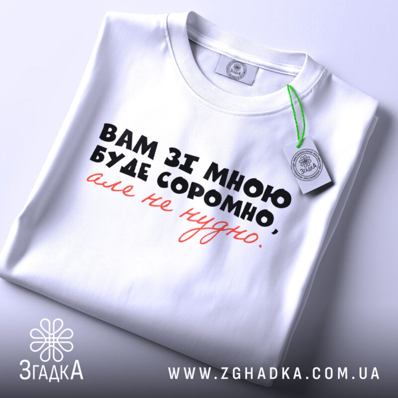 Футболка вам зі мною буде соромно. Біла футболка, складена на столі, з принтом на передній частині, де написано: 'Вам зі мною буде соромно, але не потрібно.' Принт чорного та червоного кольорів. На футболці є етикетка з зеленим шнурком. Фон світло-фіолетовий, що підкреслює чистоту дизайну.
