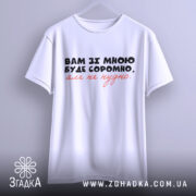 Футболка вам зі мною буде соромно. Біла футболка, підвішена на вішалці, з коротким рукавом, з принтом на грудях, де написано: 'Вам зі мною буде соромно, але не потрібно.' Принт виконаний у чорному та червоному кольорах. Фон світло-фіолетовий, що створює легкий та повітряний вигляд.