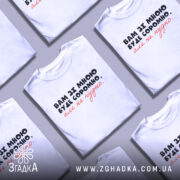 Футболка вам зі мною буде соромно. Біла футболка на чоловікові, з коротким рукавом, з принтом на грудях, де написано: 'Вам зі мною буде соромно, але не потрібно.' Принт виконаний у чорному та червоному кольорах. Фон світло-фіолетовий, а футболка виглядає стильно та сучасно.