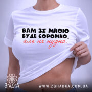 Футболка вам зі мною буде соромно. Біла футболка, складена на столі, з принтом на передній частині, де написано: 'Вам зі мною буде соромно, але не потрібно.' Принт чорного та червоного кольорів. На футболці є етикетка з зеленим шнурком. Фон світло-фіолетовий, що підкреслює чистоту дизайну.