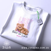 Футболка ведмедик і удача, біла, зображена на плоскій поверхні, фон світлий, принт з ведмедиком у сонцезахисних окулярах та написами 'Lucky You' і 'Best of Luck', видна етикетка.