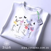 Футболка Весна Літо Осінь Зима, біла футболка, складена на поверхні, з принтом, що містить квіти та метеликів, ракурс зверху, фон світло-фіолетовий, деталі принта включають слова 'Spring', 'Summer', 'Fall', 'Winter' та 'Love for all seasons'.