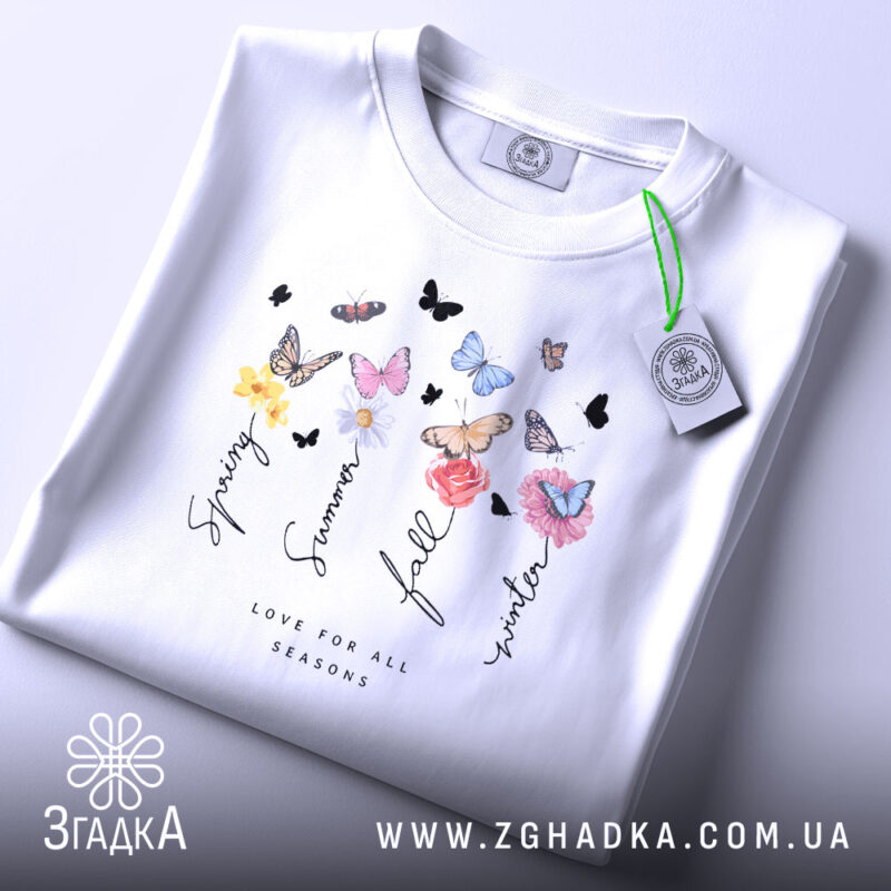 Футболка Весна Літо Осінь Зима, біла футболка, складена на поверхні, з принтом, що містить квіти та метеликів, ракурс зверху, фон світло-фіолетовий, деталі принта включають слова 'Spring', 'Summer', 'Fall', 'Winter' та 'Love for all seasons'.