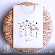 Футболка Весна Літо Осінь Зима, біла футболка, складена на плетеній кошику, з принтом, що містить квіти та метеликів, ракурс зверху, фон світло-фіолетовий, деталі принта включають слова 'Spring', 'Summer', 'Fall', 'Winter' та 'Love for all seasons'.