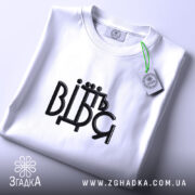 Футболка від'їбіться, складена біла футболка на світлому фоні, ракурс зверху, принт чорного кольору з написом 'ВІДЬМ', на футболці є етикетка з зеленим шнурком.