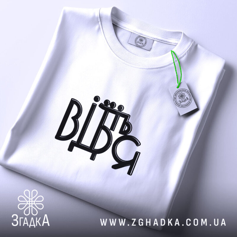 Футболка від'їбіться, складена біла футболка на світлому фоні, ракурс зверху, принт чорного кольору з написом 'ВІДЬМ', на футболці є етикетка з зеленим шнурком.