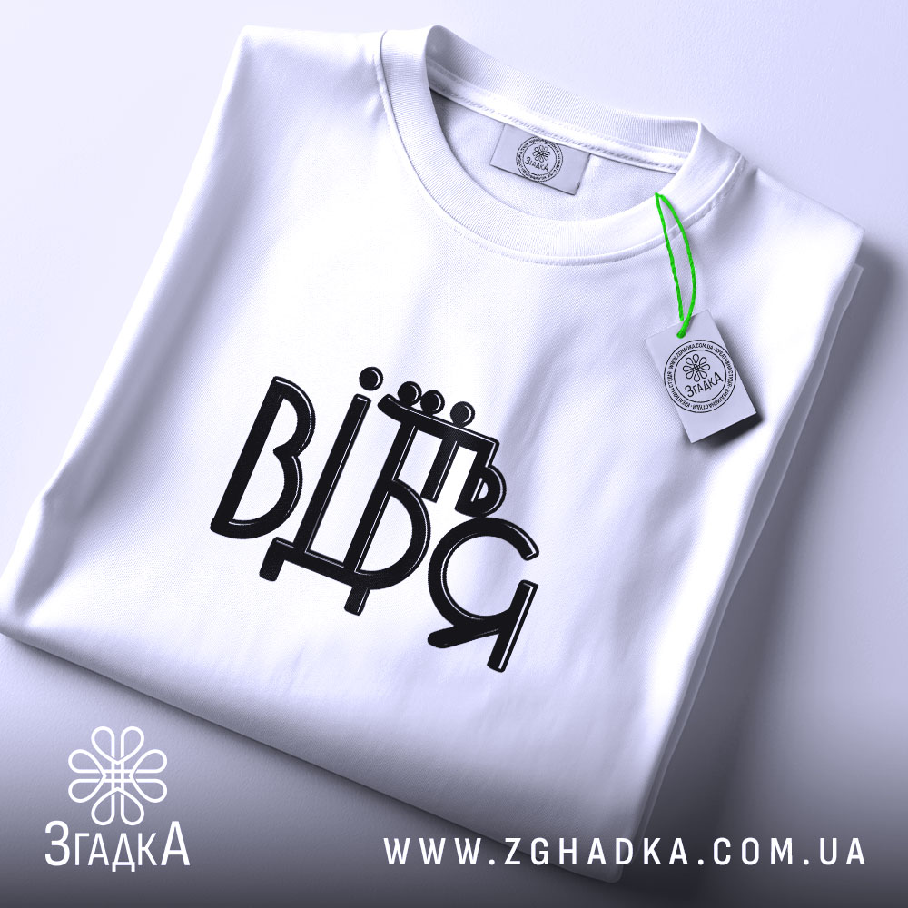 Футболка від'їбіться, складена біла футболка на світлому фоні, ракурс зверху, принт чорного кольору з написом 'ВІДЬМ', на футболці є етикетка з зеленим шнурком.