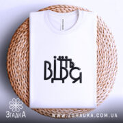 Футболка від'їбіться, складена біла футболка на плетеній кошику, ракурс зверху, фон світлий, принт чорного кольору з написом 'ВІДЬМ'.