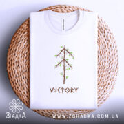 Футболка VIKTORY білого кольору, складена на круглому плетеному кошику, з принтом у вигляді дерева з коричневими гілками та зеленим листям. Фон світло-сірий, що контрастує з текстурою кошика.