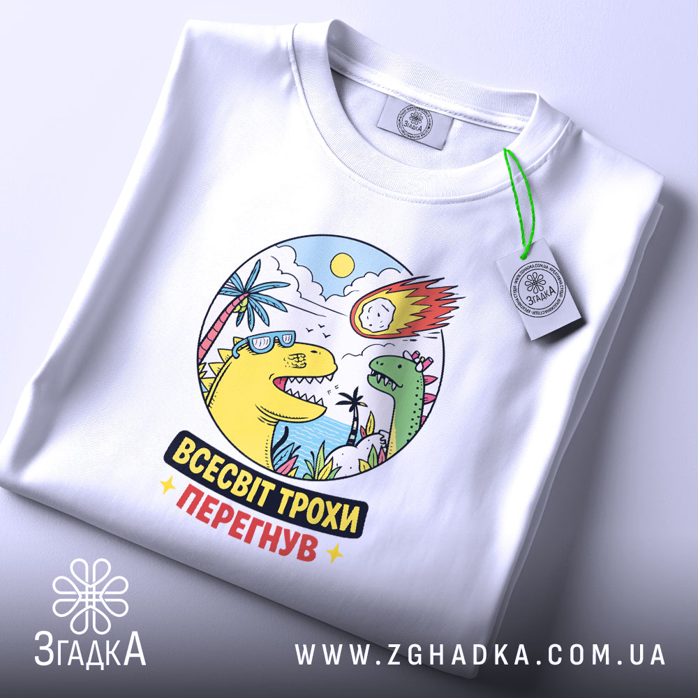 Футболка всесвіт трохи перегнув, біла, на плоскій поверхні, ракурс зверху, фон світло-фіолетовий, принт з динозаврами, сонцем і метеоритом, яскраві кольори, текст "ВСЕСВІТ ТРОХИ ПЕРЕГНУВ", етикетка на рукаві.