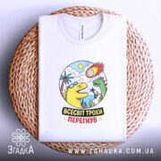 Футболка всесвіт трохи перегнув, біла, на плоскій поверхні, ракурс зверху, фон світло-фіолетовий, принт з динозаврами, сонцем і метеоритом, яскраві кольори, текст "ВСЕСВІТ ТРОХИ ПЕРЕГНУВ", футболка на плетеній основі.